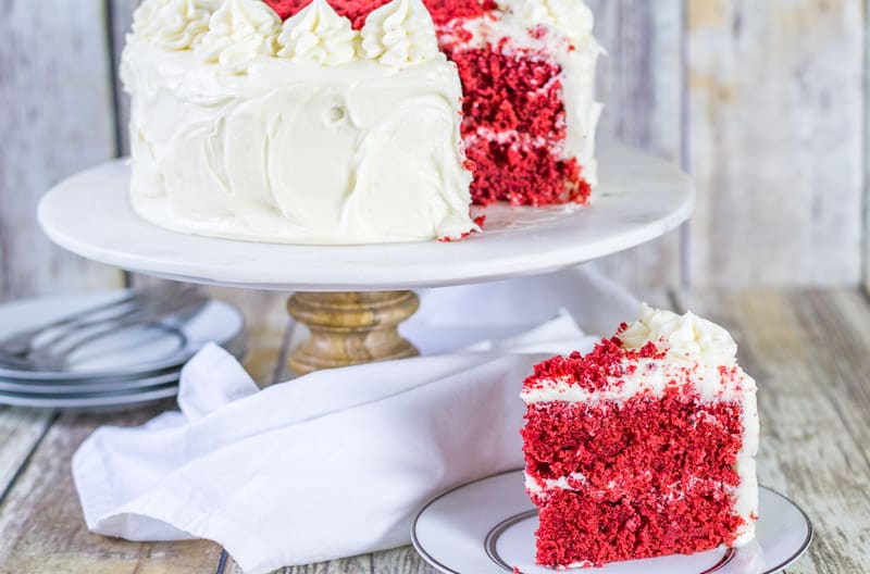 Sour Cream Red Velvet Cake on a marble cake stand with slice on a white plate