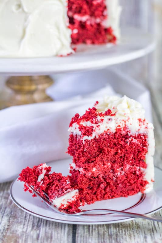 Slice of Sour Cream Red Velvet Cake on a white plate with a fork with cake in the background