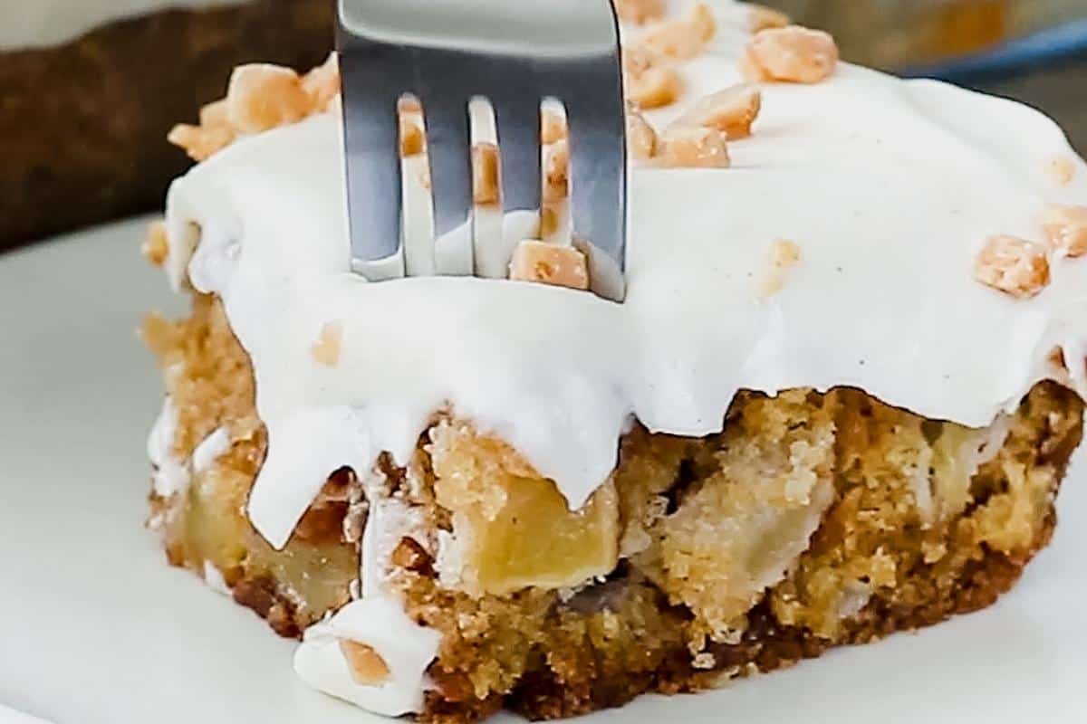 Close up of fork cutting into cake with icing and apple chunks.