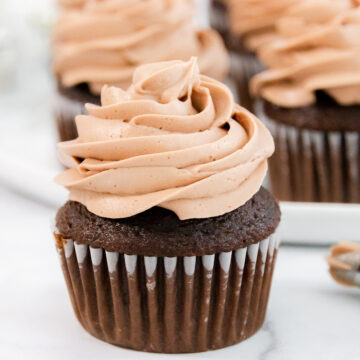 Piped chocolate frosting on a cupcake with a piping bag next to it and cupcakes behind it.
