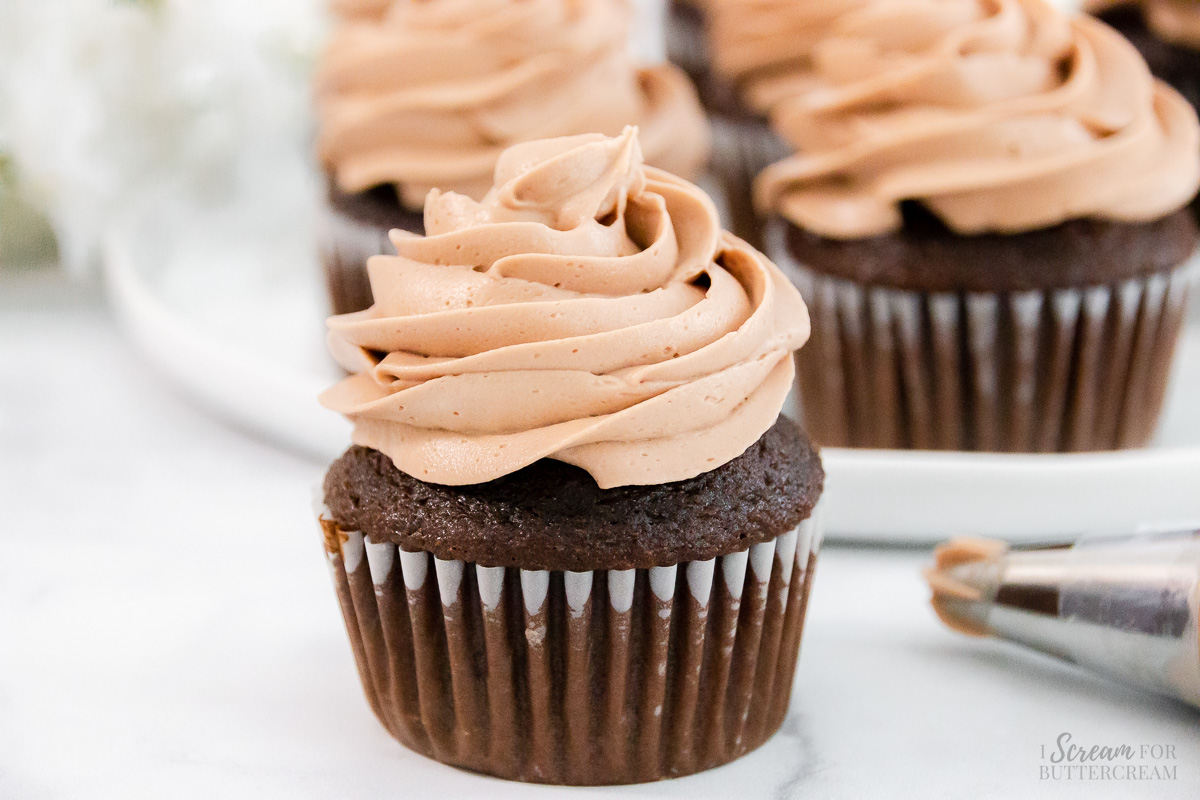 Piped chocolate frosting on a cupcake with a piping bag next to it and cupcakes behind it.