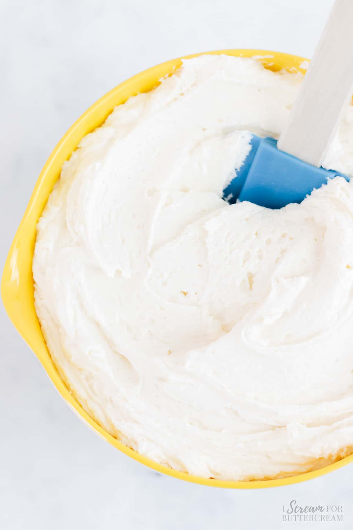 Vanilla buttercream frosting in a yellow bowl with a blue spatula.