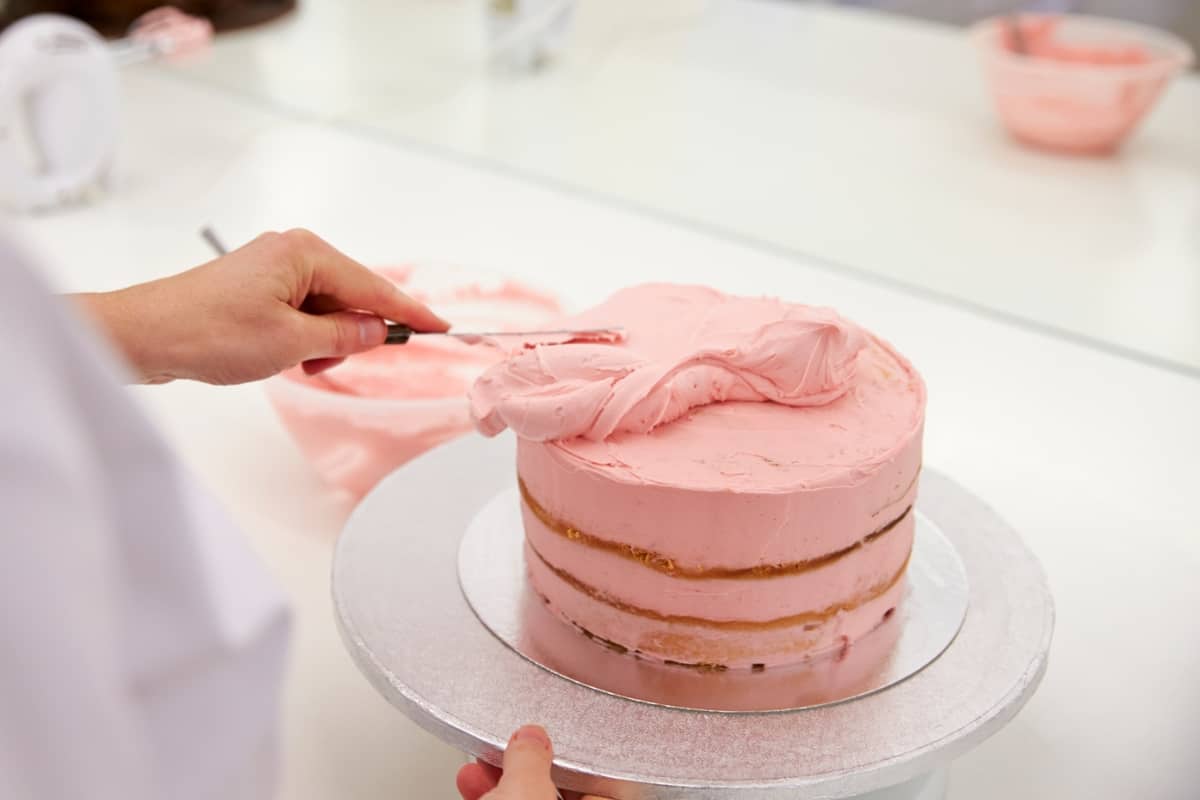 A woman icing a pink cake on a white table.