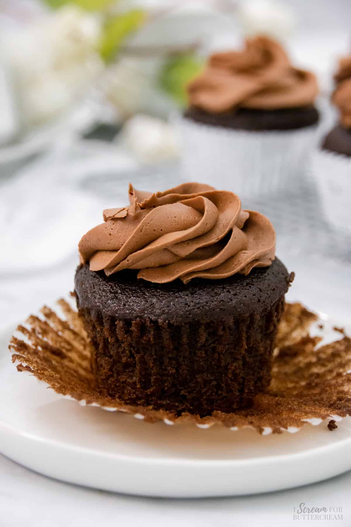 One chocolate cupcake unwrapped with chocolate frosting on top on a white plate.
