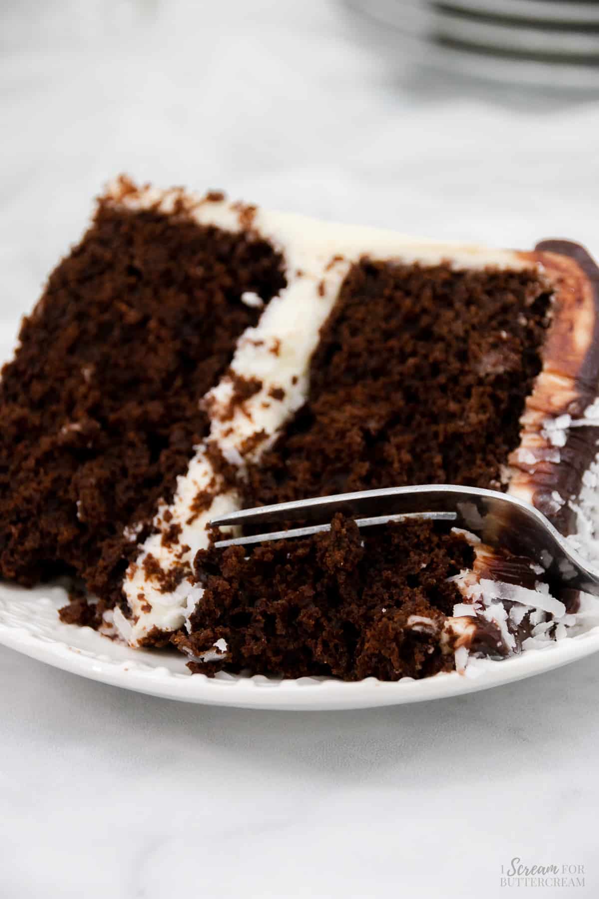 Close up image of coconut flavored chocolate cake with white frosting with a fork on a white plate.