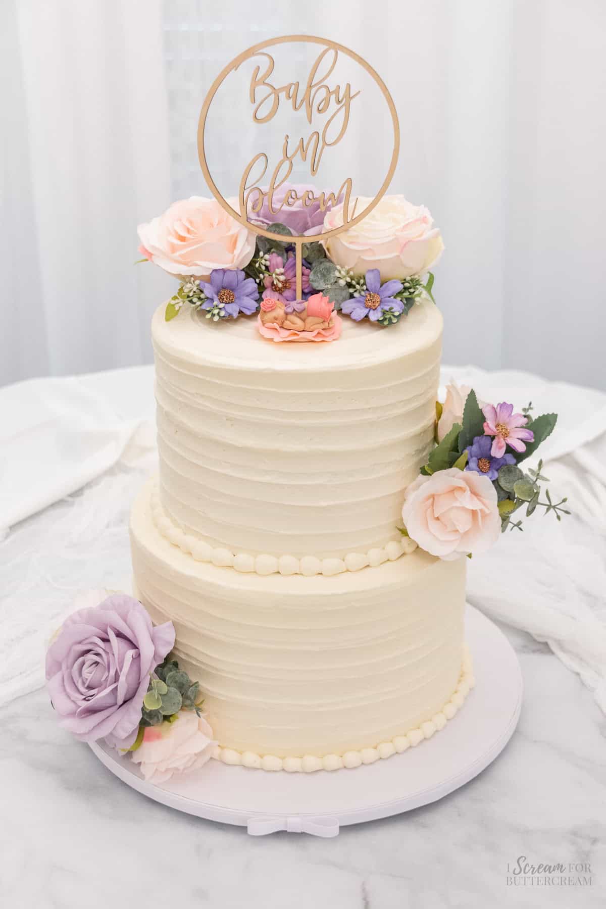 Baby in bloom decorated baby shower cake for a girl with vanilla buttercream and two cake tiers with purple and pink flowers, a fondant baby on top and a wooden cake topper that says baby in bloom.