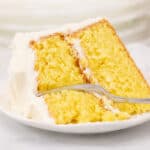 Wide image of slice of lemon sour cream cake on a white plate with frosting and a fork.