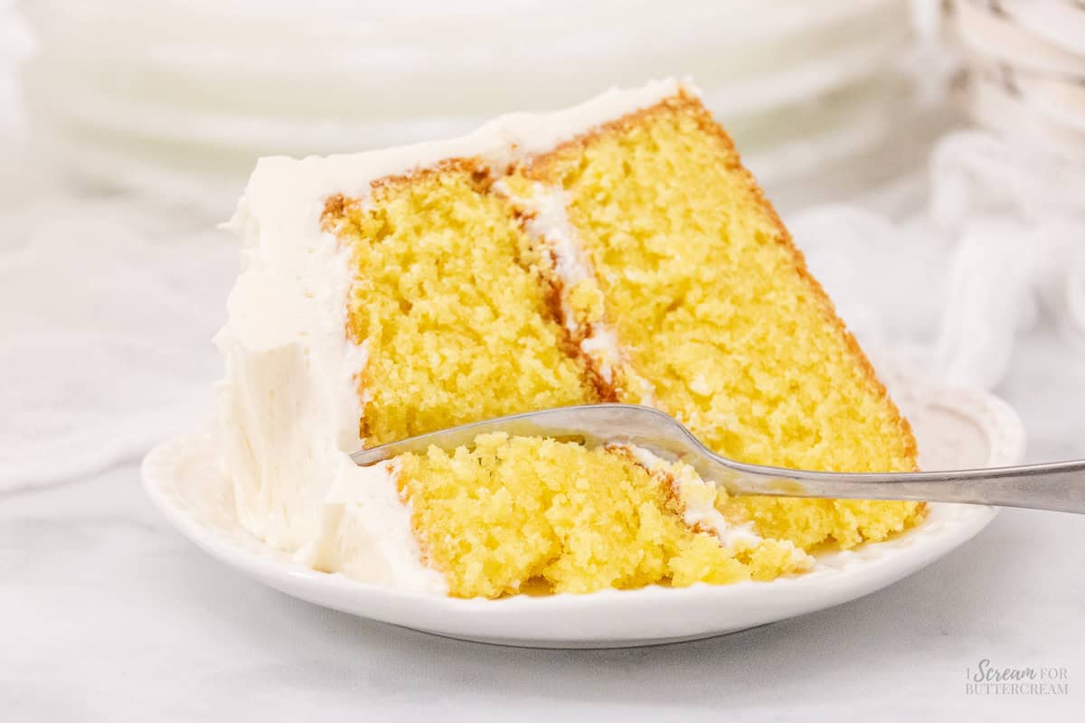 Wide image of slice of lemon sour cream cake on a white plate with frosting and a fork.