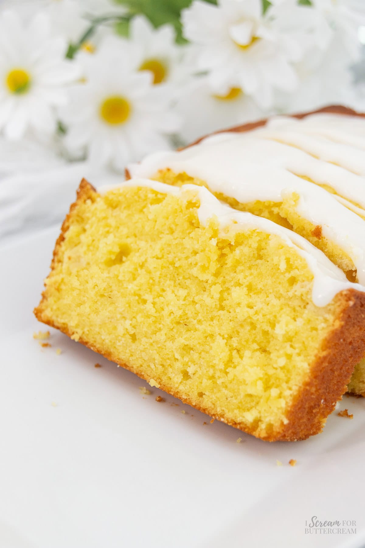 Sliced lemon pound cake on a white platter with glaze on top and daisies in the background.