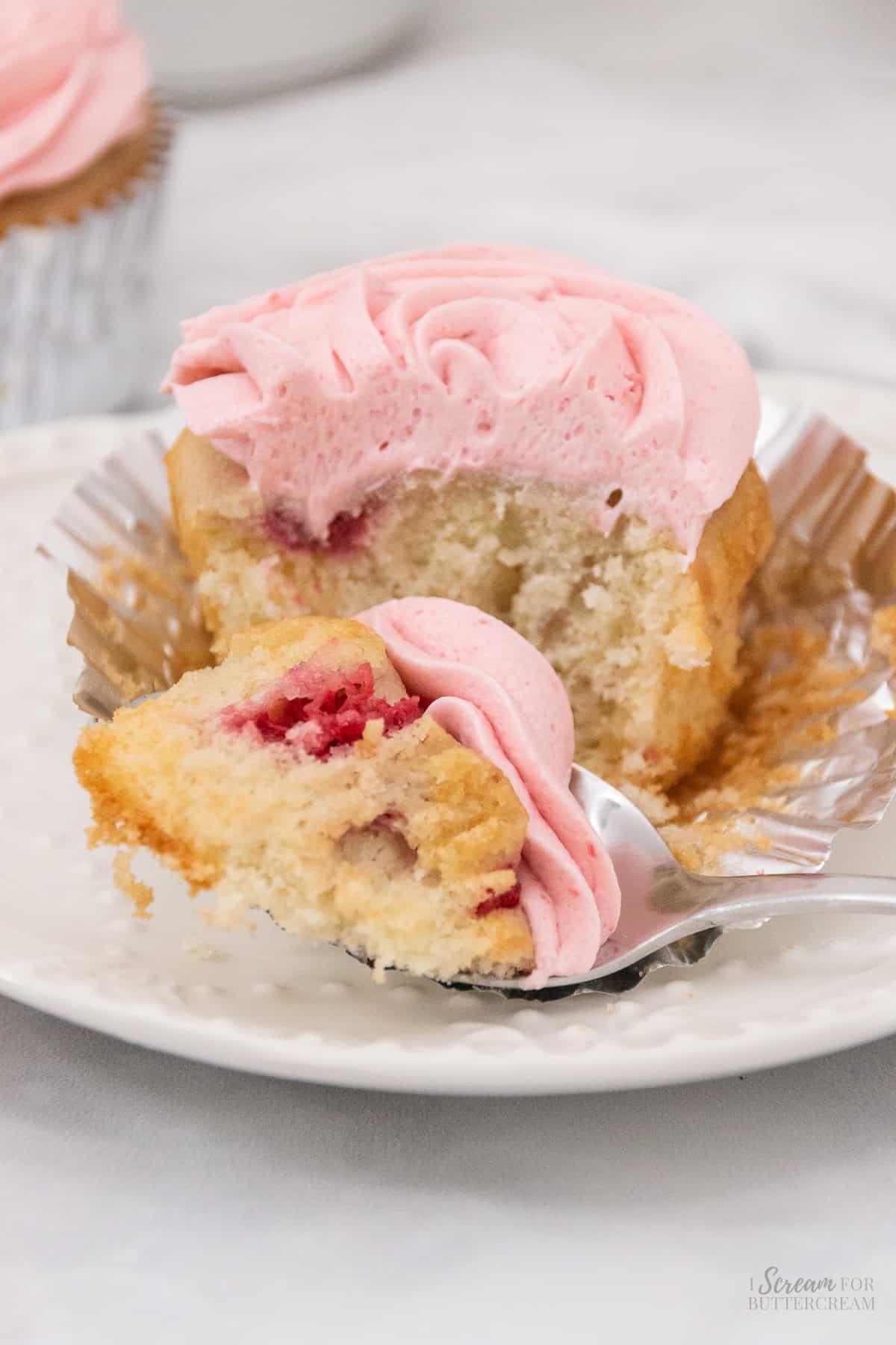 Close up image of a raspberry vanilla cupcake with frosting on a white plate with a fork and a small piece of the cupcake on the fork.