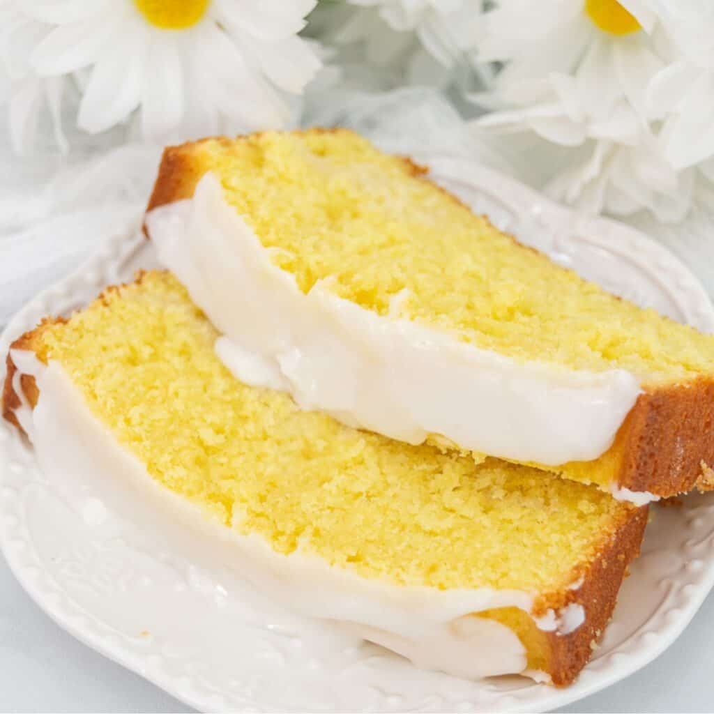 Sliced lemon pound cake on a white plate with glaze on top and daisies in the background.