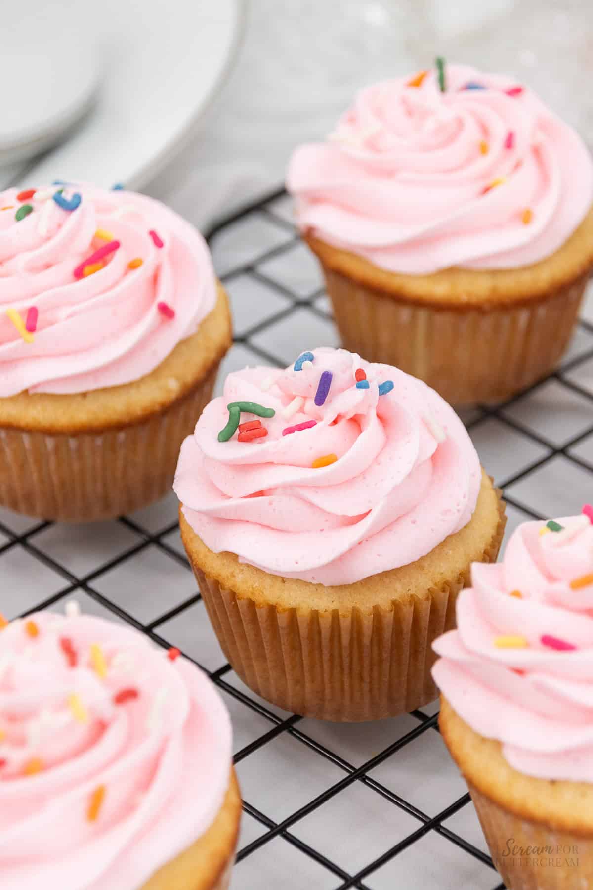 Top view of vanilla cupcakes with pink frosting and sprinkles on a cooling rack.