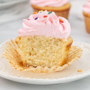 Wide image of cut vanilla sour cream cupcake with frosting and sprinkle son a white plate.