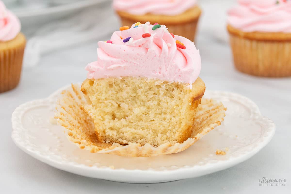 Wide image of cut vanilla sour cream cupcake with frosting and sprinkle son a white plate.