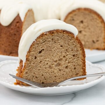 The image shows a slice of a bundt cake with a soft, moist texture, topped with a generous layer of creamy white frosting. The cake has a light brown color, indicating it may contain spices. The background features the rest of the bundt cake with the frosting elegantly dripping down the sides. The slice is placed on a white plate, and a fork rests beside it, ready for serving. The cake has a classic homemade appearance, evoking a comforting, autumnal feel.