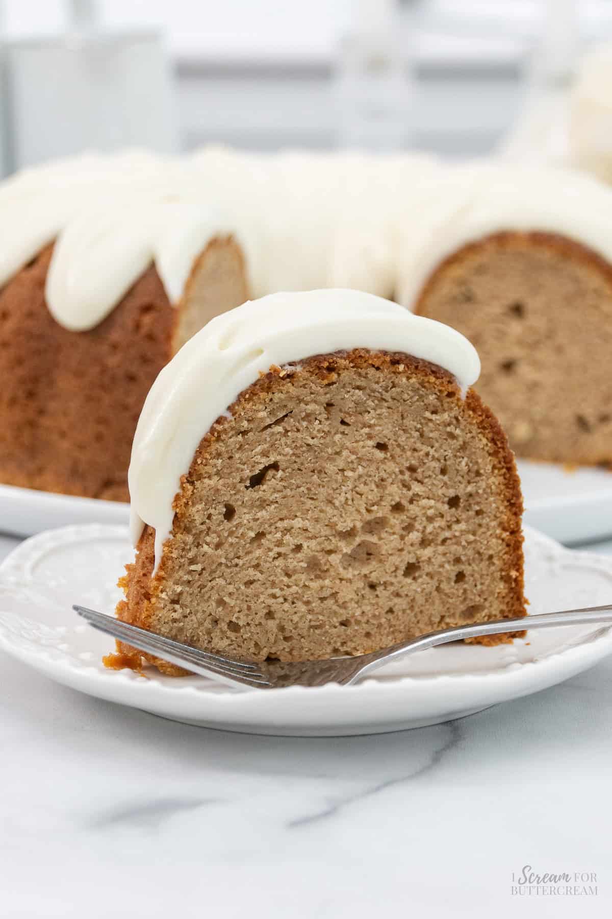 A slice of moist, spiced bundt cake with creamy white frosting sits on a white plate with a fork, showcasing its soft texture and inviting flavor.