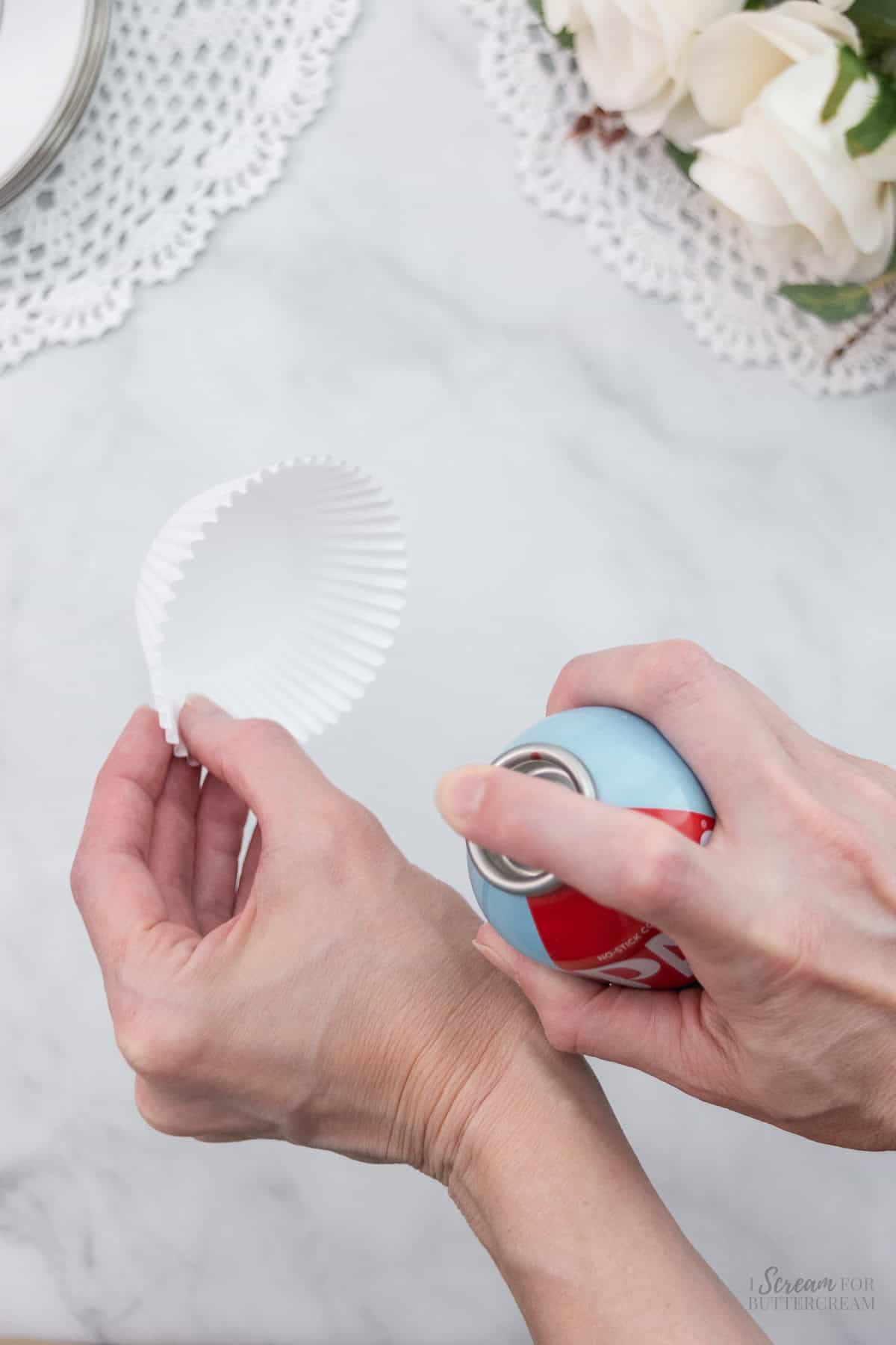 A hand holds a white cupcake liner while spraying it with nonstick cooking spray, with white roses and lace decor in the background.