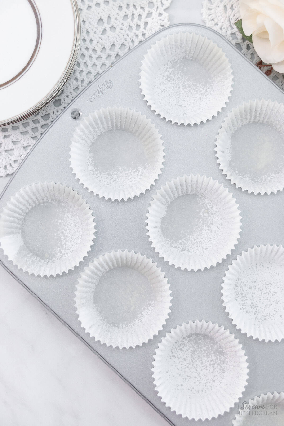 A clean cupcake pan lined with white paper liners that have been lightly sprayed with nonstick cooking spray, with decorative plates and lace in the background.