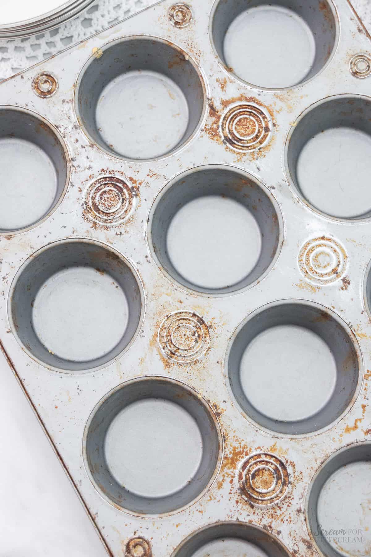 A used cupcake pan showing signs of rust and baked-on stains, with worn spots in several of the cups.