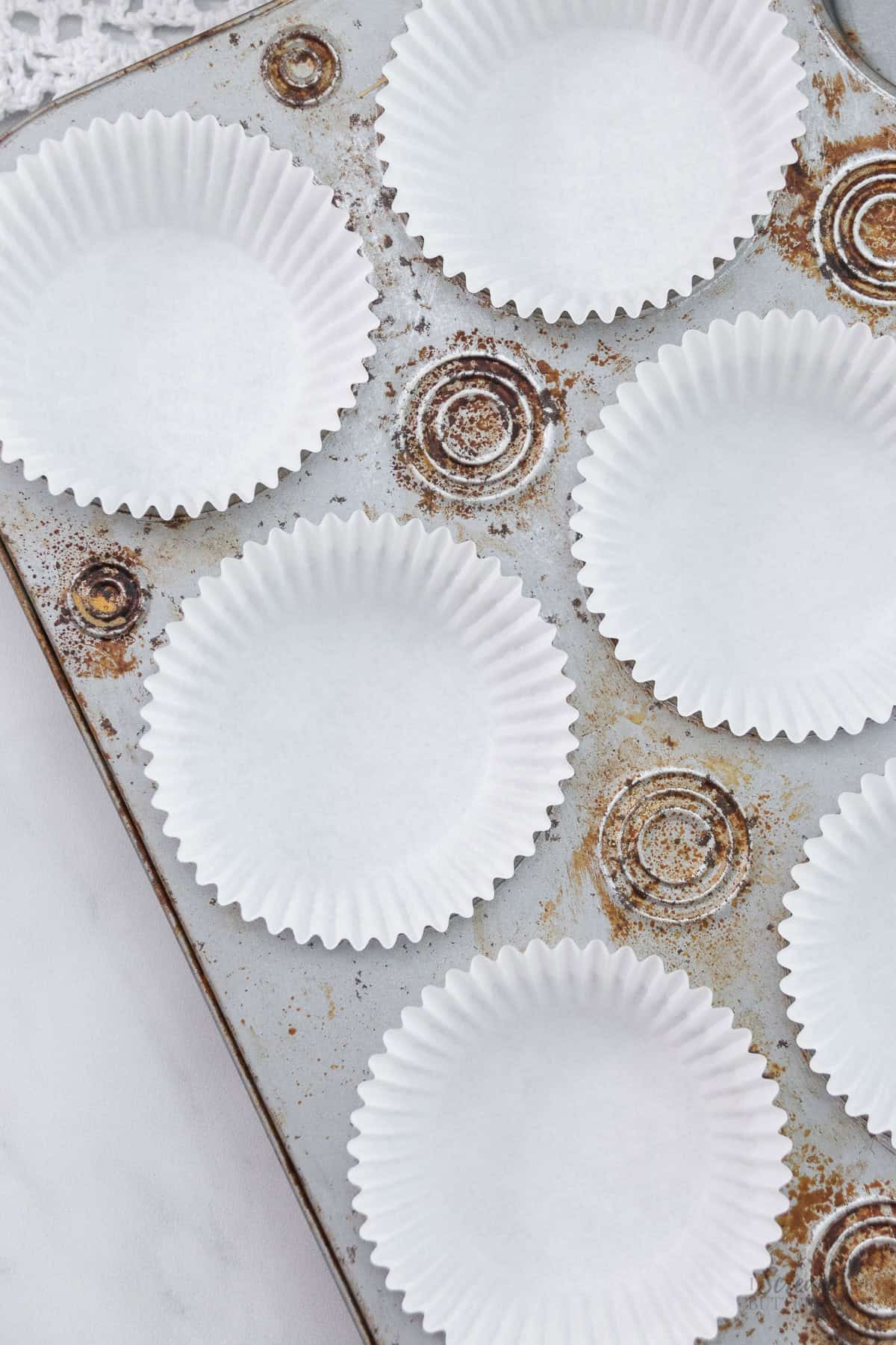 A used cupcake pan with rust and baked-on stains, lined with clean white paper cupcake liners.