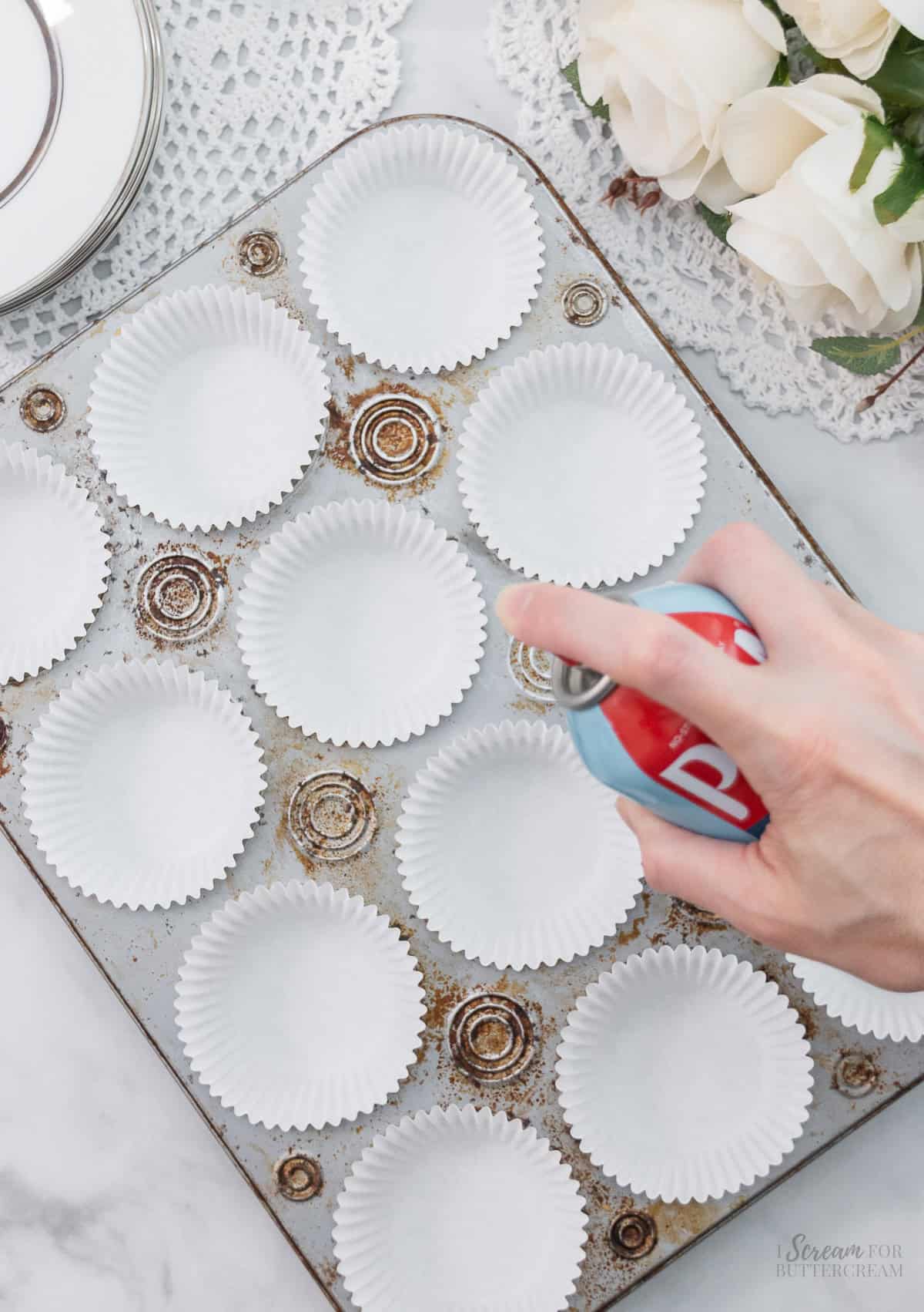 A hand sprays nonstick cooking spray into white paper liners arranged in a rust-stained cupcake pan, with white roses in the background.