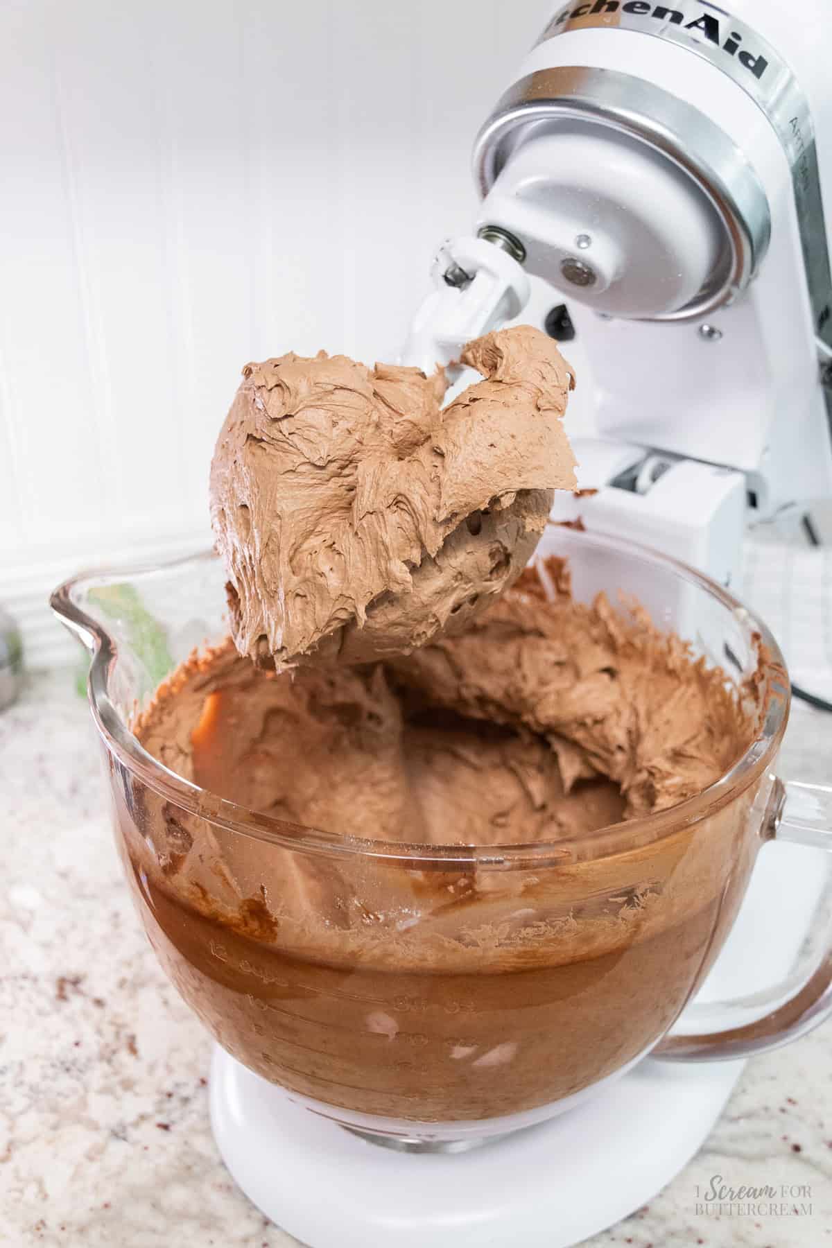 A stand mixer with a paddle attachment covered in creamy chocolate buttercream frosting, with a glass mixing bowl filled with more frosting on the countertop.