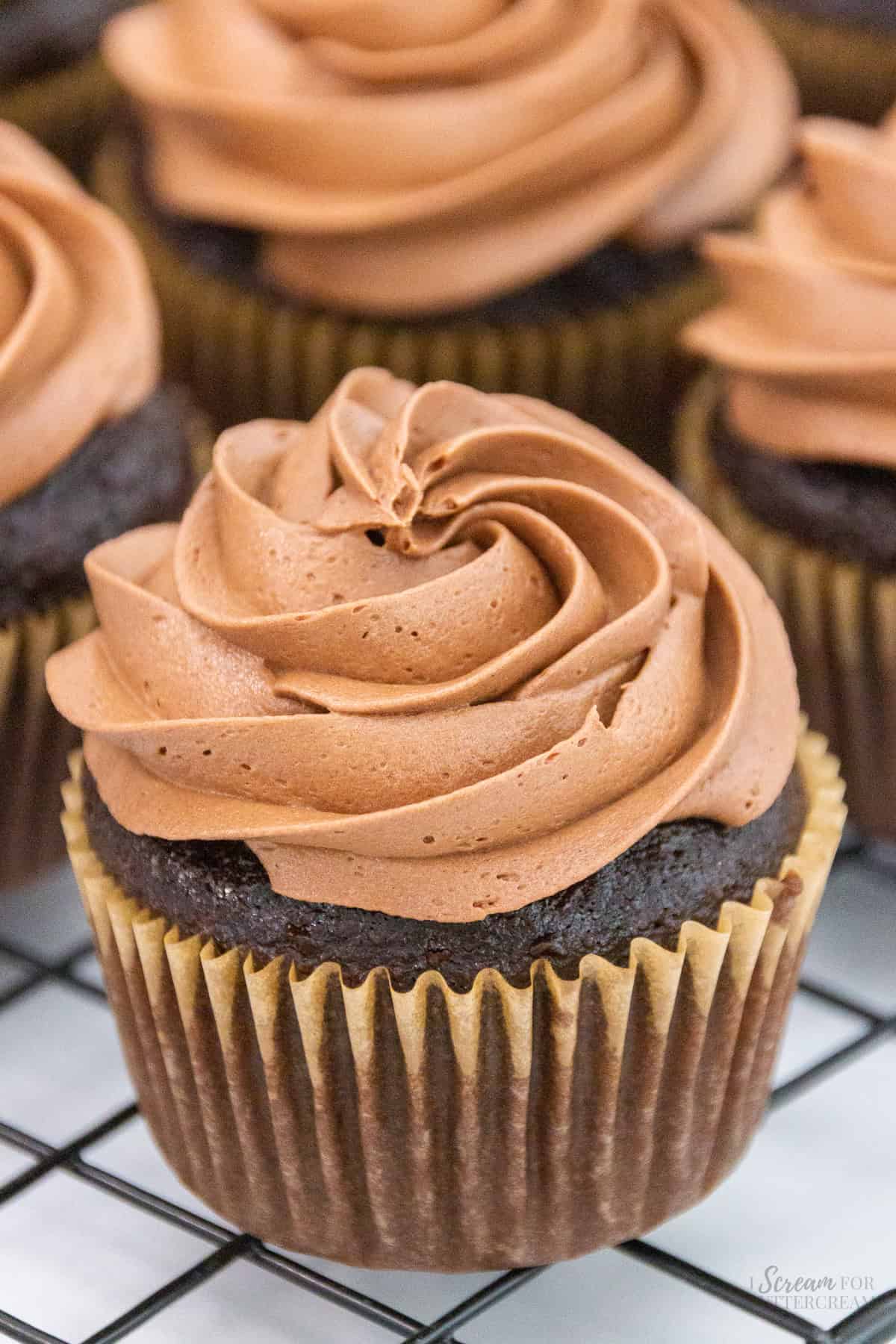 A chocolate cupcake with a swirl of rich chocolate buttercream frosting, surrounded by other frosted cupcakes on a cooling rack.