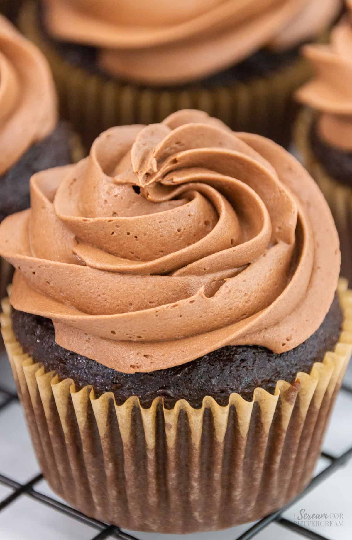 A close-up of a chocolate cupcake topped with a smooth swirl of rich chocolate buttercream frosting, resting on a cooling rack.