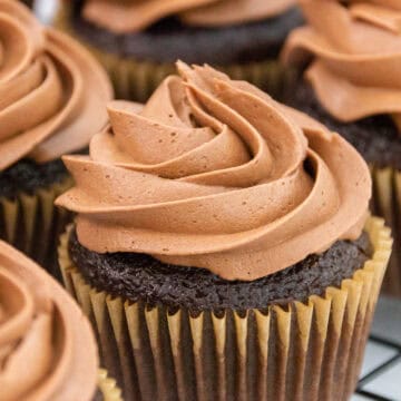 A group of chocolate cupcakes topped with swirls of rich chocolate buttercream frosting, arranged on a cooling rack.