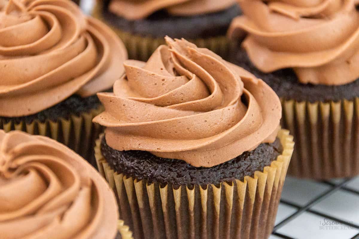 A group of chocolate cupcakes topped with swirls of rich chocolate buttercream frosting, arranged on a cooling rack.