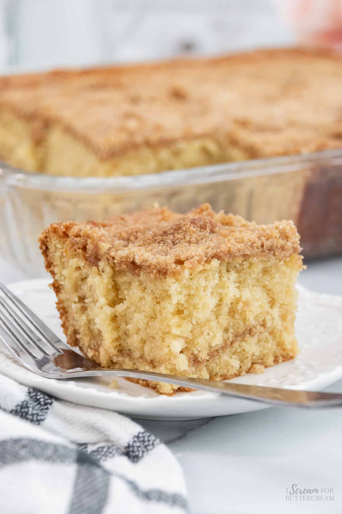 A slice of buttermilk coffee cake with a crumbly cinnamon-sugar topping on a white plate with a fork, with the rest of the cake in a glass baking dish in the background.