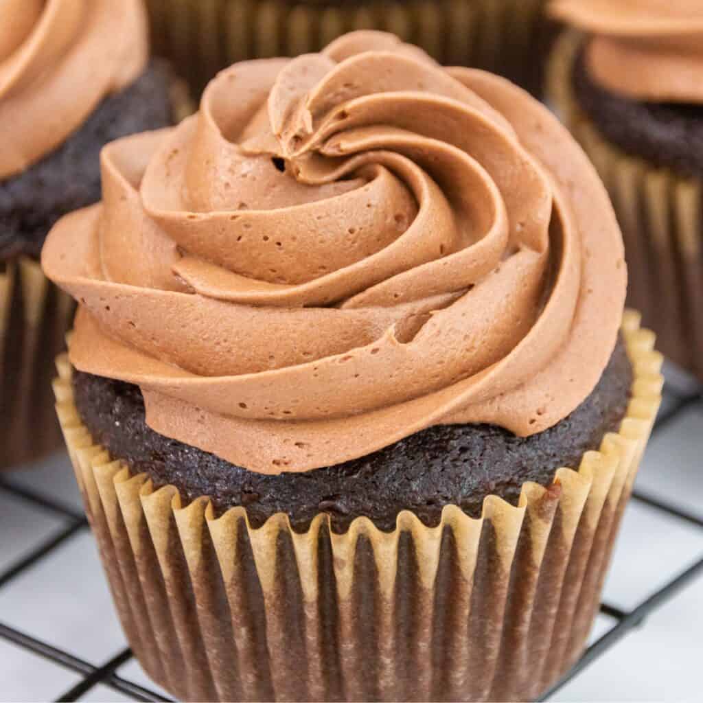 A close-up of a chocolate cupcake topped with a swirl of rich chocolate buttercream frosting on a cooling rack.