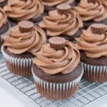 A batch of chocolate cupcakes with chocolate peanut butter buttercream frosting and peanut butter cups on top, arranged on a cooling rack.