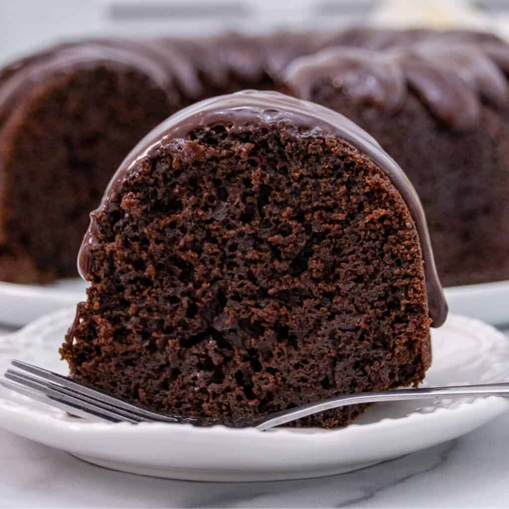 A slice of rich chocolate bundt cake with glossy chocolate glaze on a white plate, accompanied by the rest of the cake in the background.