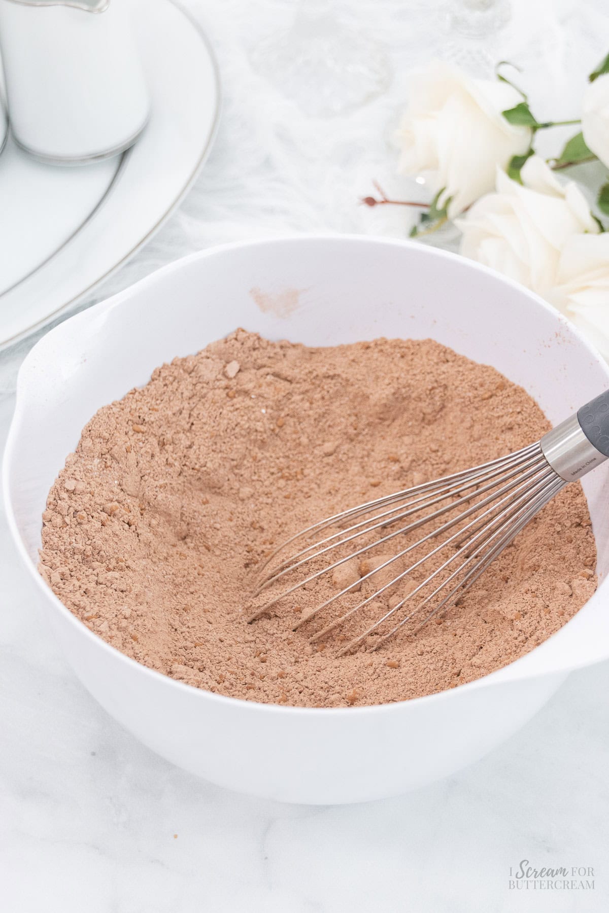 A white mixing bowl filled with cocoa powder-based dry ingredients, being whisked with a metal whisk, placed on a light-colored surface with soft floral decor in the background.