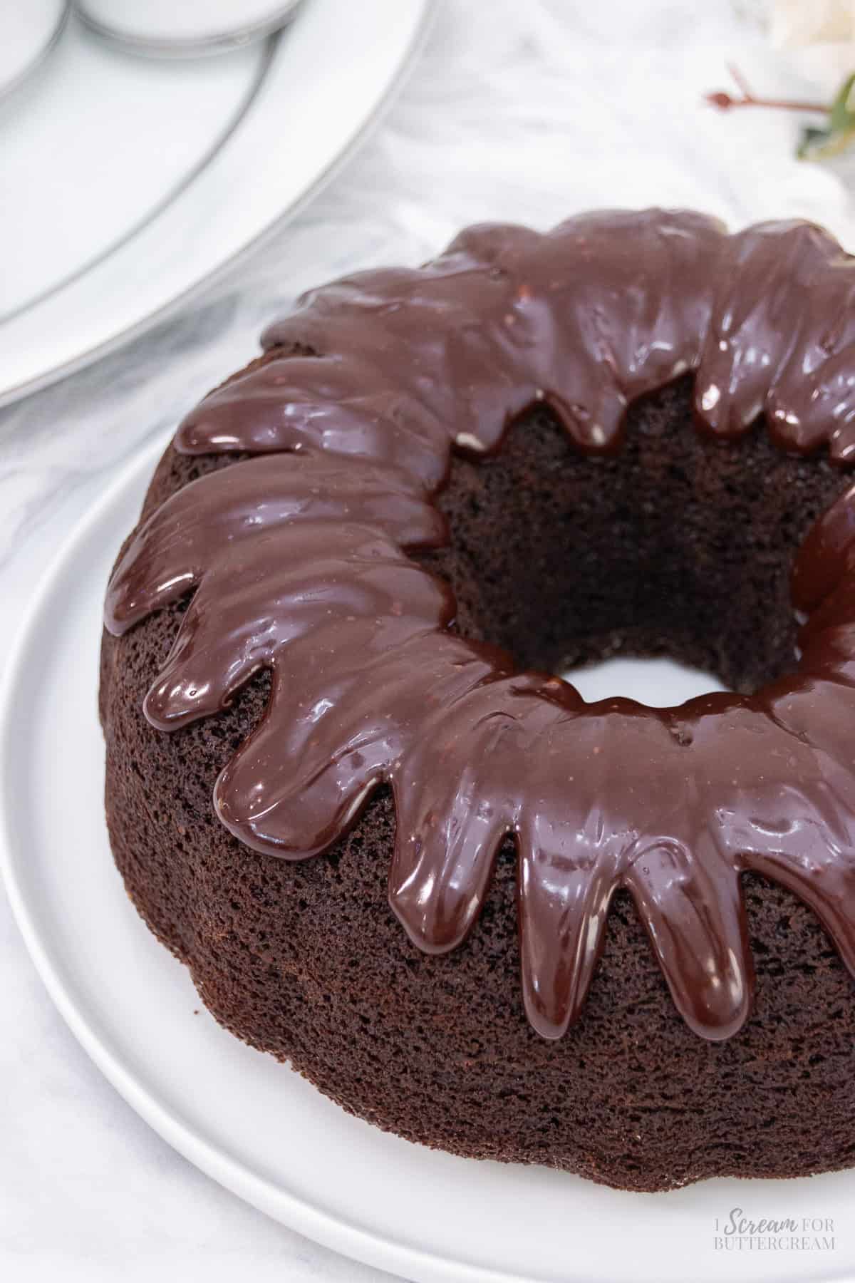 A frosted chocolate bundt cake with a glossy chocolate glaze dripping over the edges, placed on a white plate against a light background.