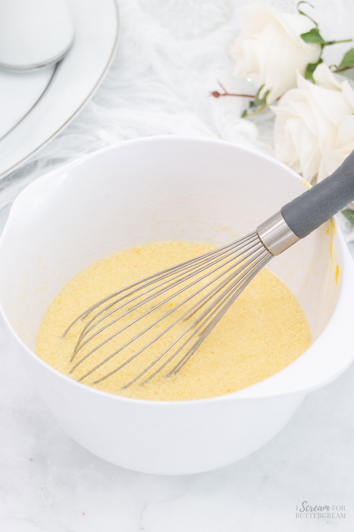 A white mixing bowl containing liquid ingredients being whisked with a metal whisk, on a light surface with soft floral accents in the background.