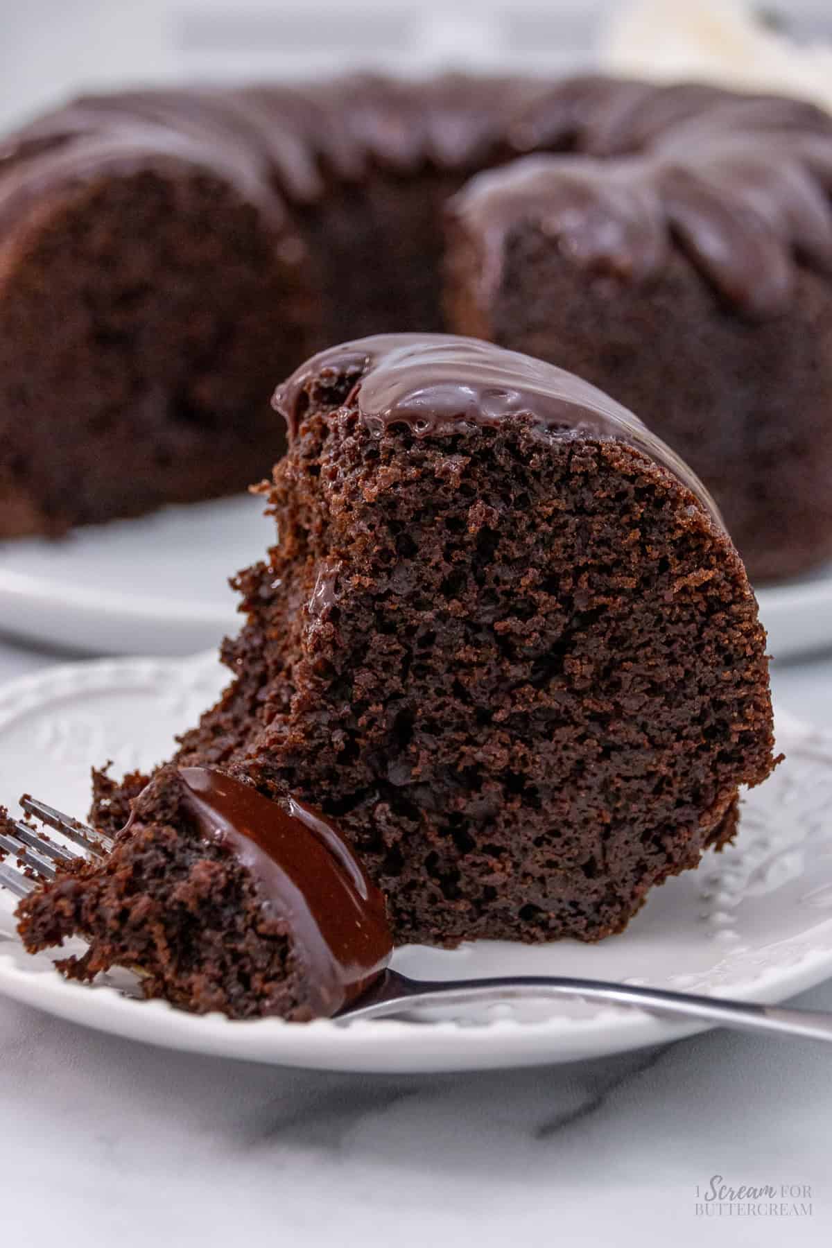 A close-up of a slice of moist chocolate bundt cake with a glossy chocolate glaze on a white plate, with a forkful of cake in the foreground and the full cake in the background.