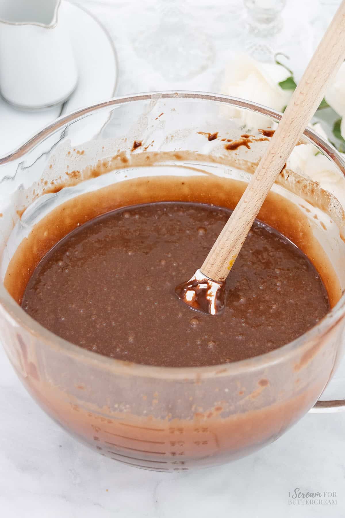 A glass mixing bowl filled with chocolate cake batter, on a light surface with soft floral decor in the background.