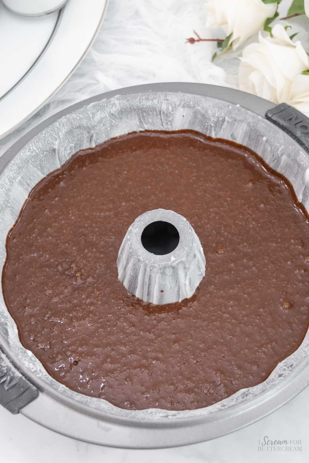 A floured bundt pan filled with smooth chocolate cake batter, ready to be baked, placed on a light surface with soft floral accents in the background.