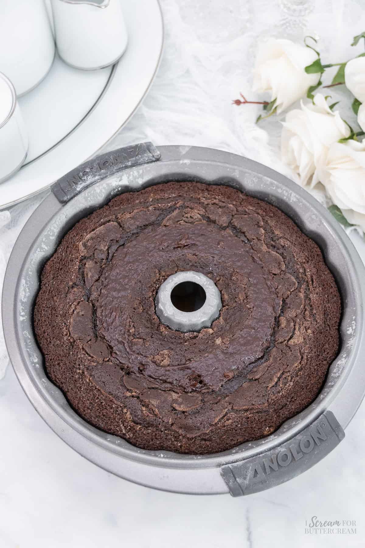 A freshly baked chocolate bundt cake cooling in the pan, on a light surface with soft floral accents in the background.