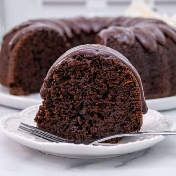 A slice of rich chocolate bundt cake with glossy chocolate glaze on a white plate, accompanied by the rest of the cake in the background.