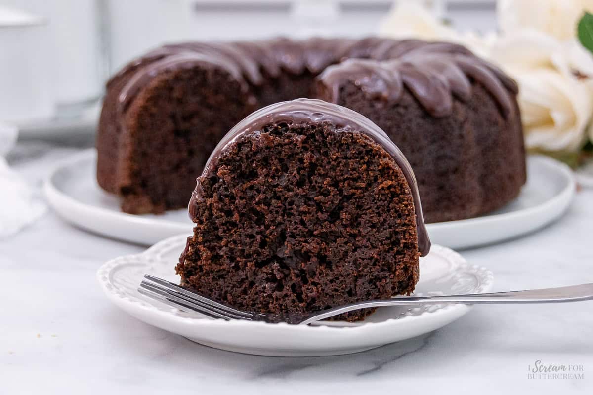 A slice of rich chocolate bundt cake with glossy chocolate glaze on a white plate, accompanied by the rest of the cake in the background.