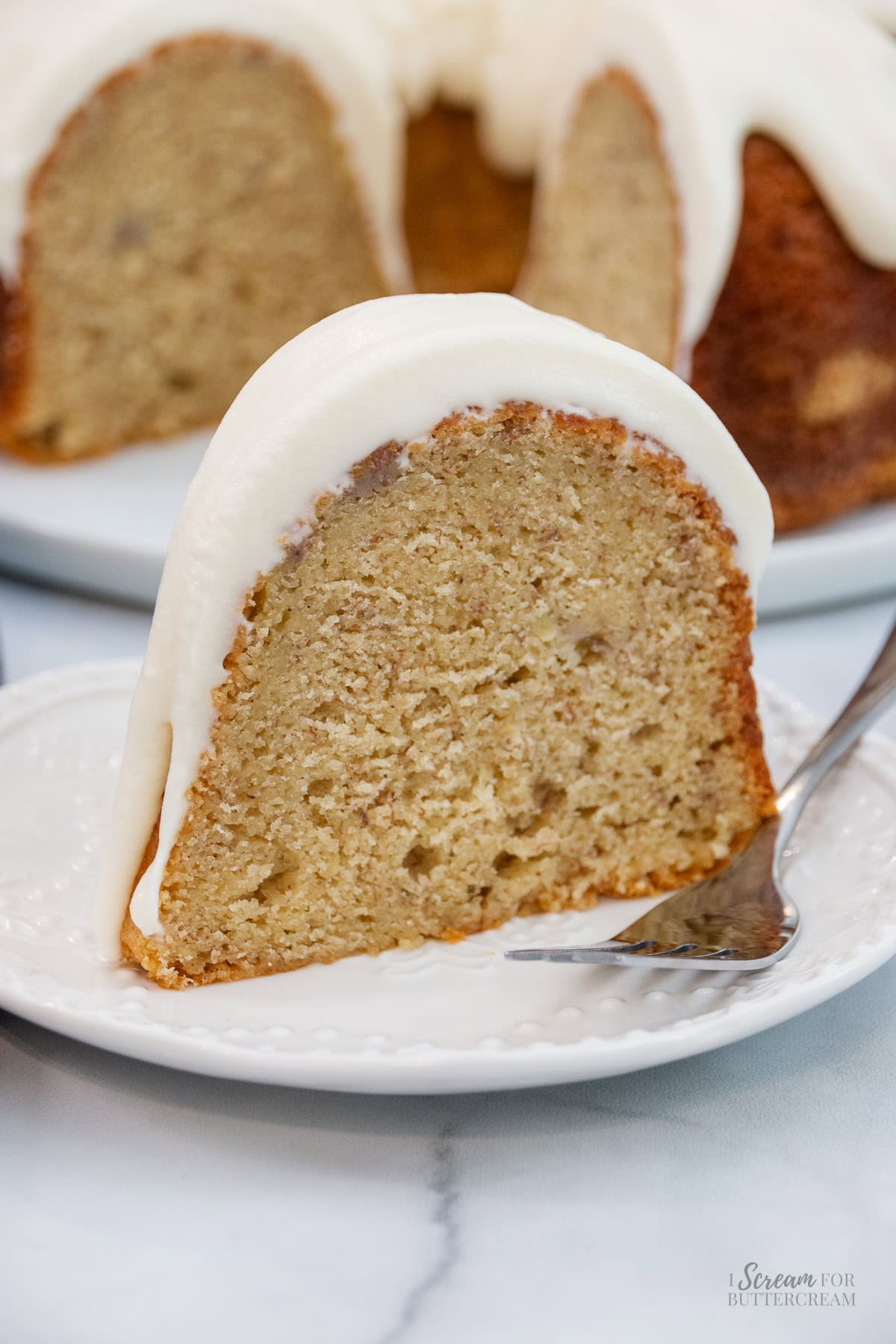 Tall image of a single thick slice of banana pound cake on a decorative white plate with a fork, showing its soft, fluffy texture and rich frosting.