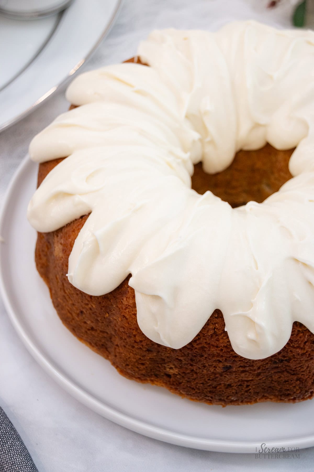 Overhead close-up of a freshly baked banana pound cake with thick, swirled cream cheese frosting generously spread on top, displayed on a white plate.