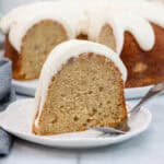 Wide shot of a slice of banana pound cake with glaze, placed on a plate in front of the full Bundt cake and surrounded by soft decor.