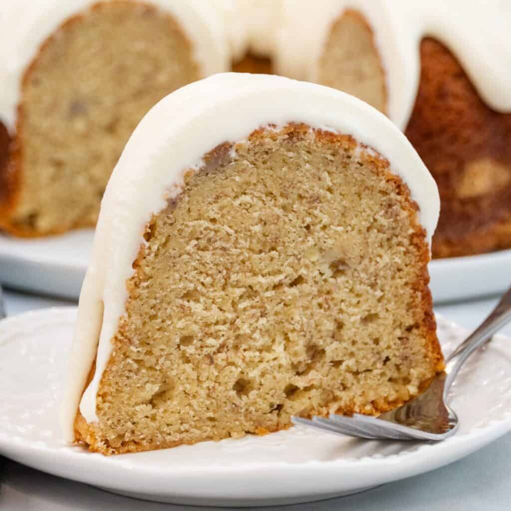 Close-up of a thick slice of moist banana pound cake topped with creamy vanilla frosting, sitting on a white plate with a fork.