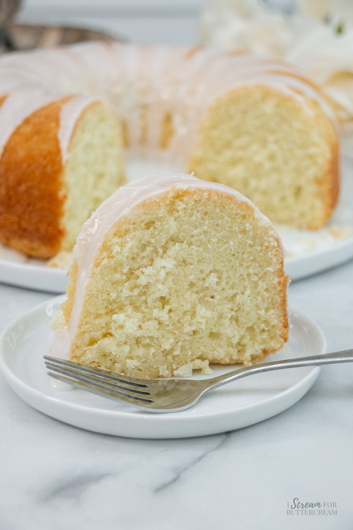 Slice of glazed coconut pound cake on a plate with a fork, full bundt cake in the background.
