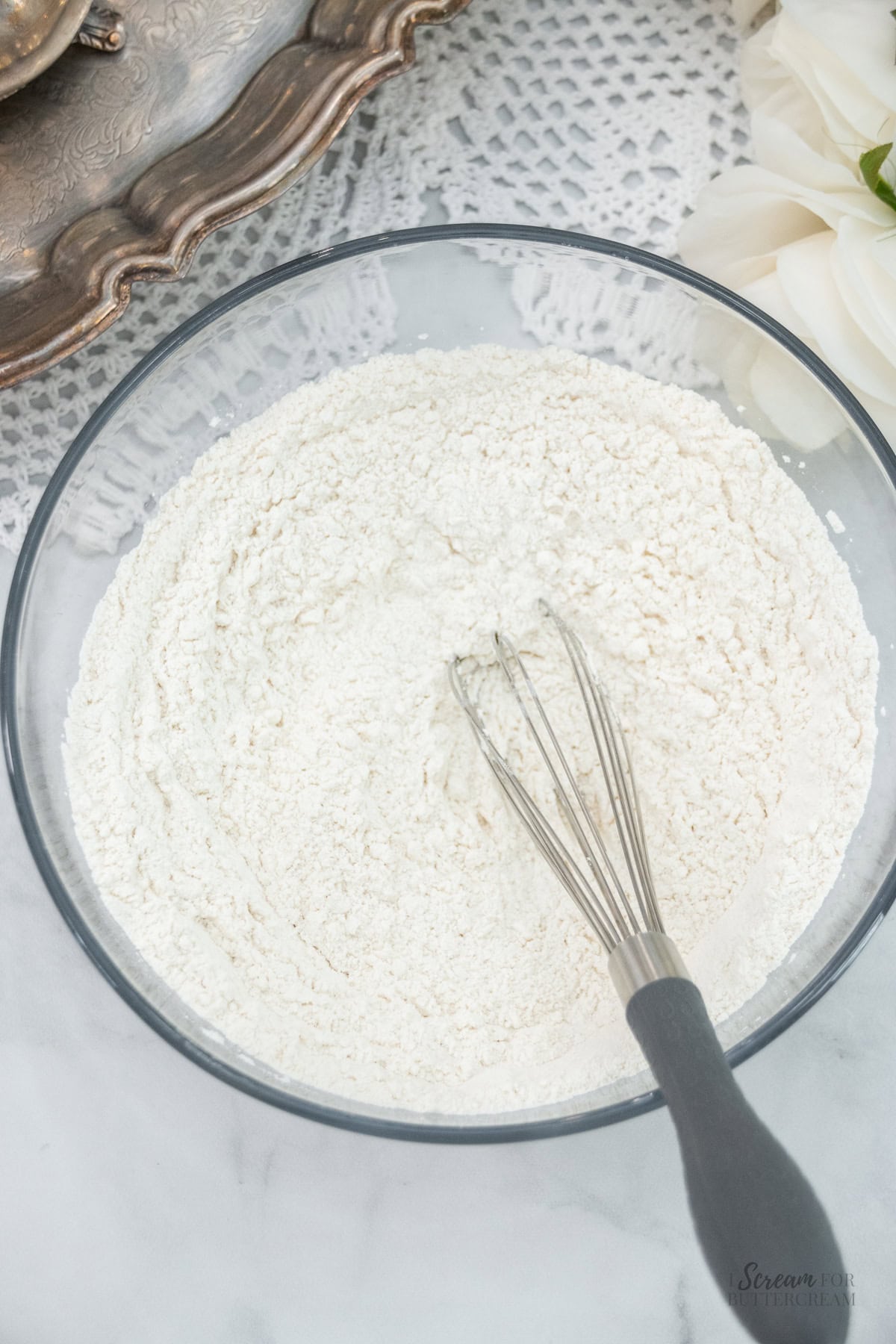 Glass bowl of dry cake ingredients with a whisk, set on a marble surface.