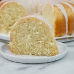 Slice of coconut pound cake on a white plate with the full bundt cake in the background.